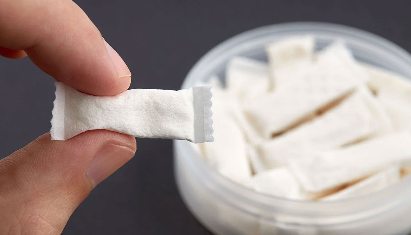 Person holding a nicotine pouch between index finger and thumb over an empty tub of pouches against a grey gradient background.